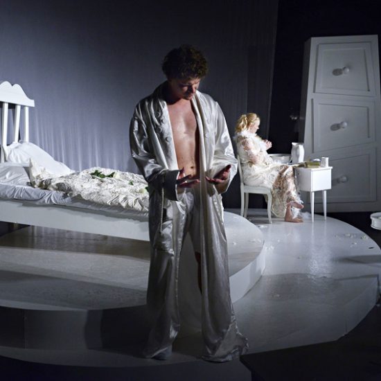 Man i silverfärgad morgonrock med en kvinna i bakgrunden i en vitt spetsigt nattlinne.