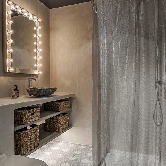 Toalett inredning i betongfärg med ett grafitgrått handfat och spegel med lampor runt sig.