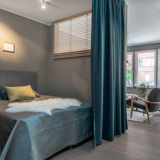Blågardin som rumsavskiljare mellan ett sovrum med dubbelsäng och vardagsrum i en kontorsliknande lägenhet