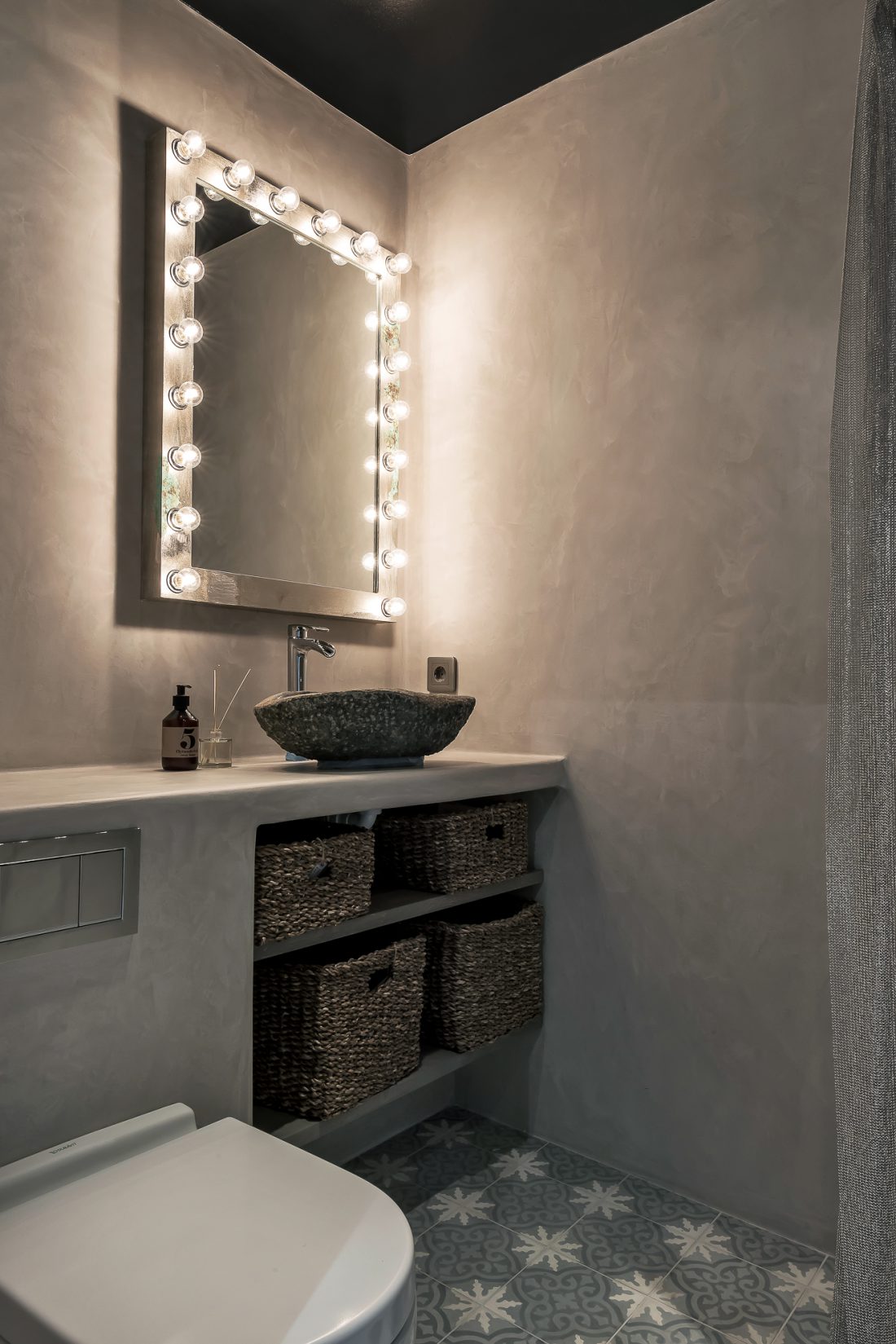 Toalett inrett i betong med en stor spegel med lampor runt sig.