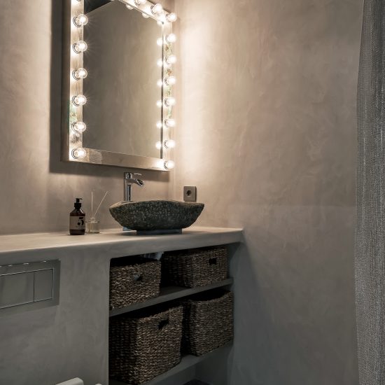 Toalett inrett i betong med en stor spegel med lampor runt sig.